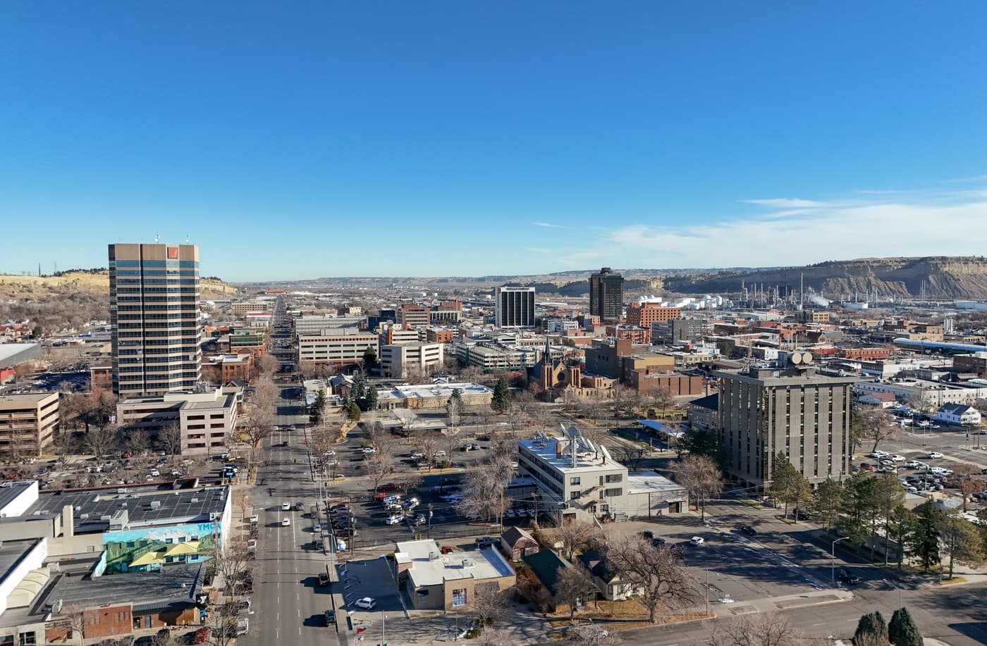 The downtown of Billings from the air under a clear blue sky: two tall office towers and a few mid-rise buildings among low blocks and parking lots, a long straight street running away at the left, and a line of pale sandstone cliffs along the horizon.