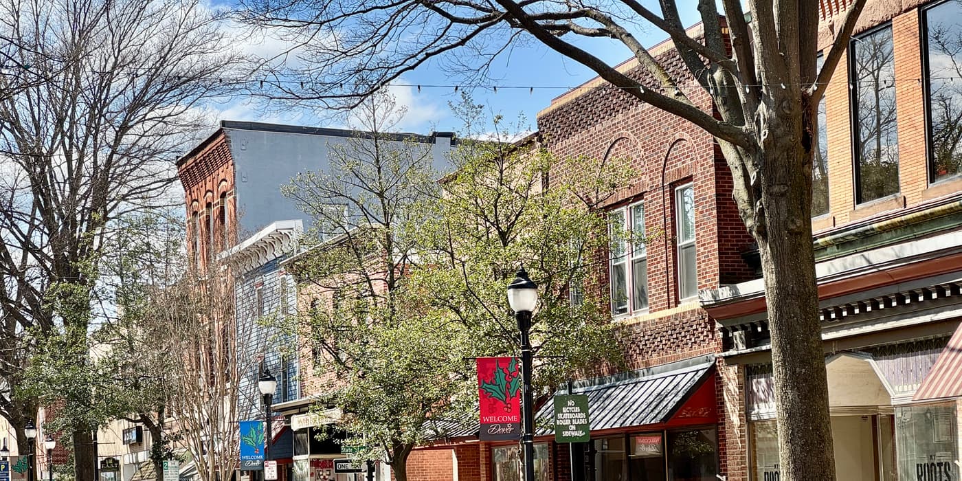 The upper floors and shopfronts of a street of two- and three-story brick buildings in bright sun, under trees coming into leaf. A black lamp post in the middle carries a red banner that reads Welcome.