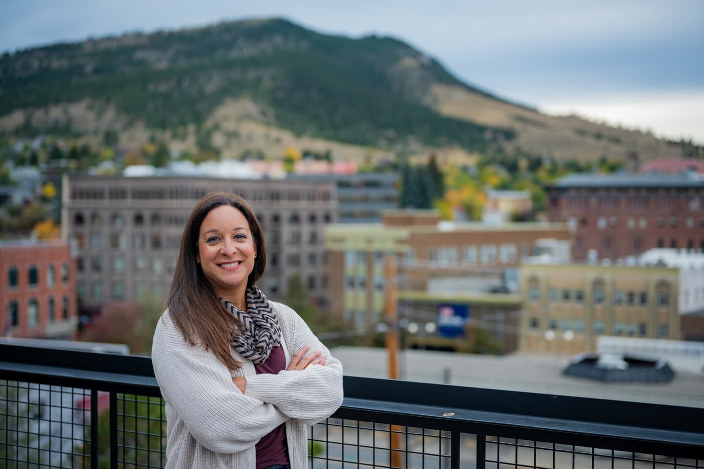Alani Bankhead, in a white cardigan and a patterned scarf, stands smiling with her arms folded at the railing of a terrace. Behind her, out of focus, are the brick buildings of a downtown and a wooded hill.