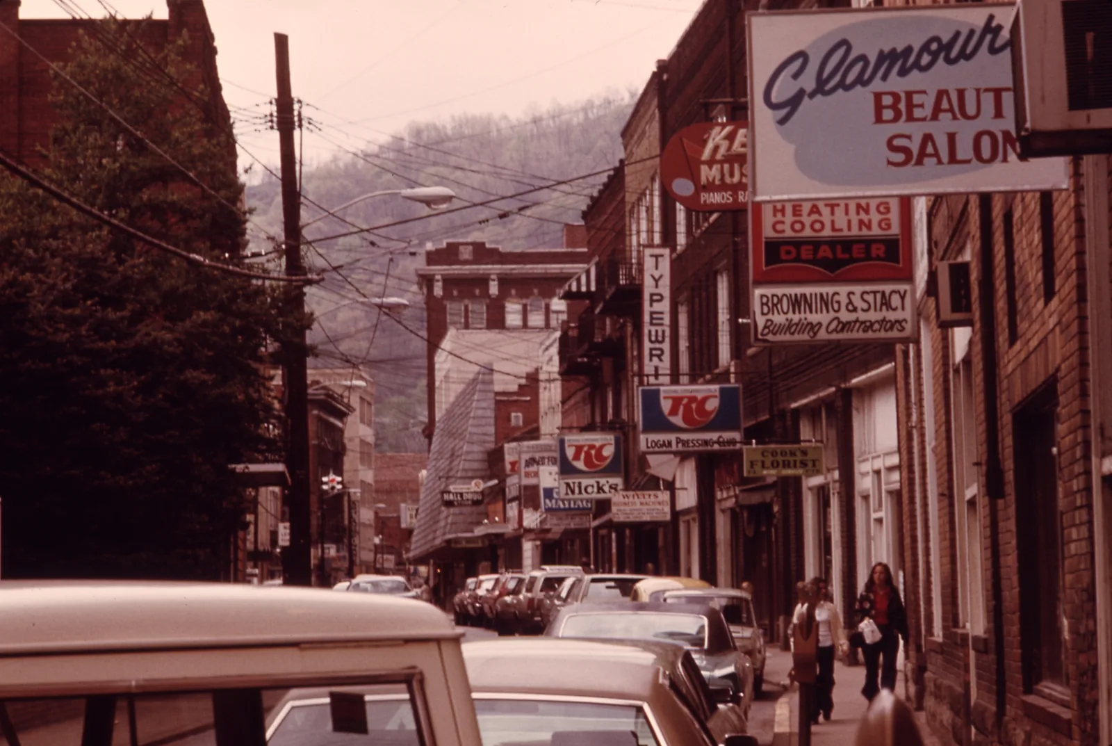 A narrow main street in a hill town in 1974: shop signs, parked cars along one side, wooded hills behind.