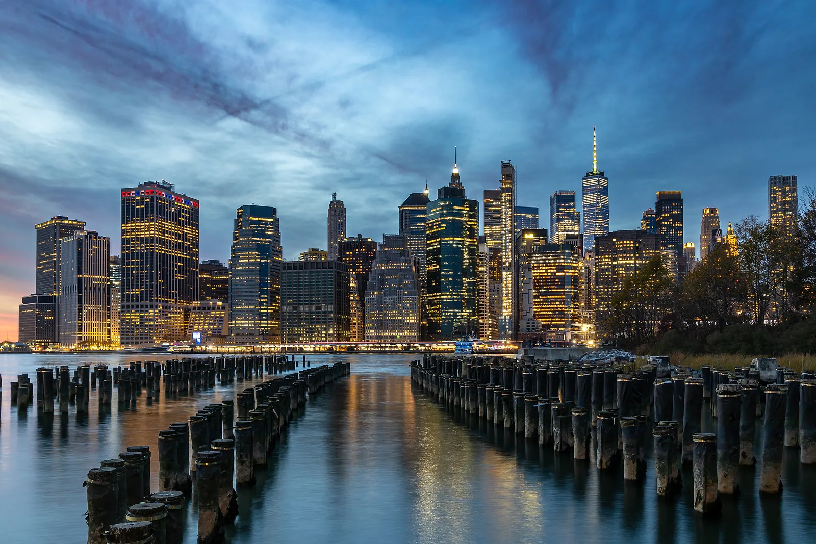 Lower Manhattan's towers lit at dusk, seen across the East River past rows of old pier pilings.