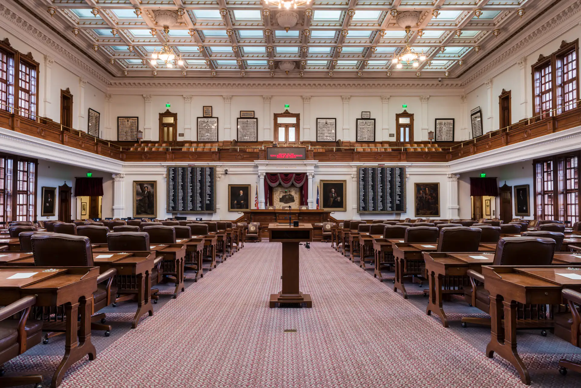 The empty chamber of the Texas House of Representatives: rows of wooden desks and leather chairs on either side of an aisle that runs to the front microphone and the speaker’s rostrum, under a coffered glass ceiling.