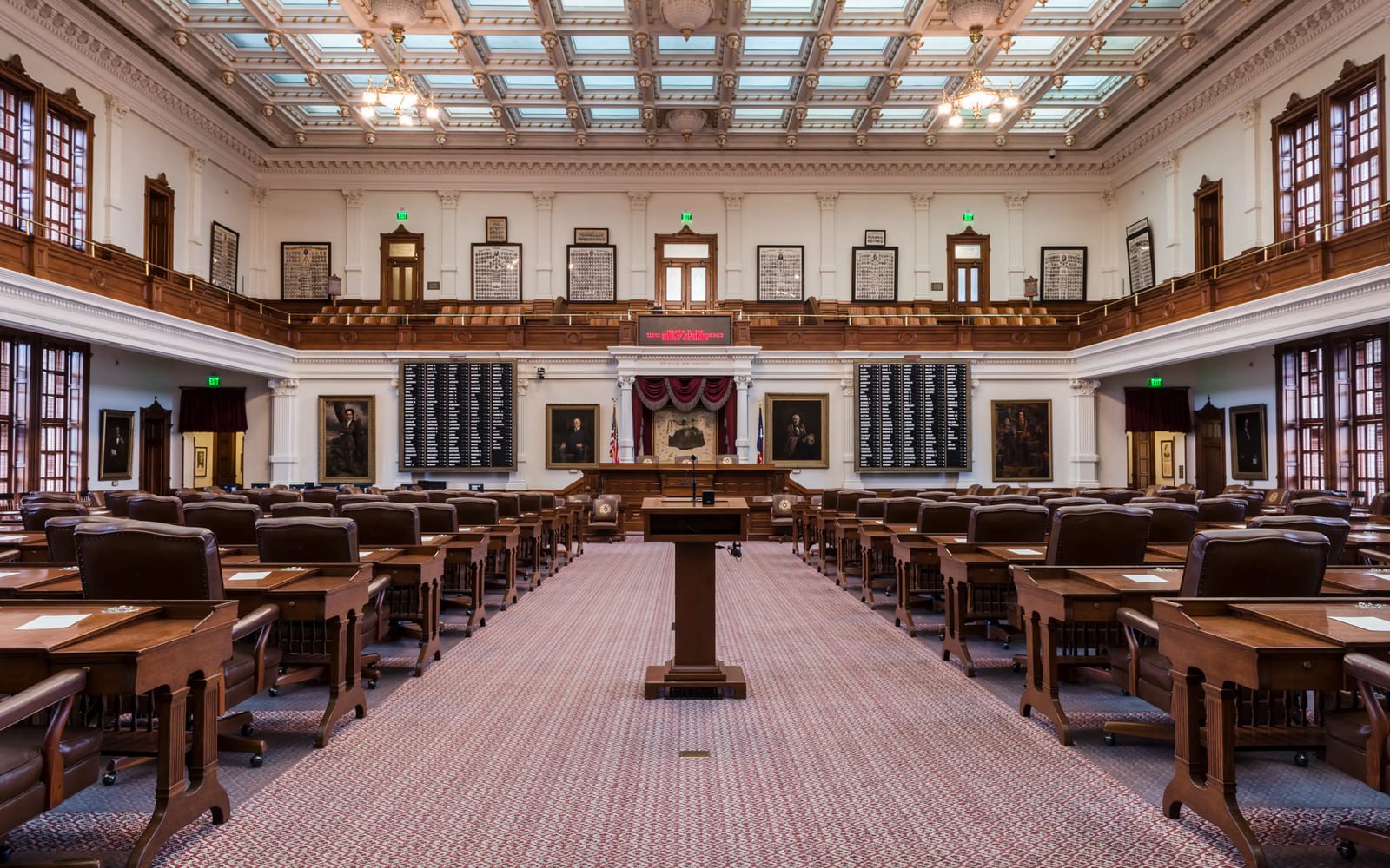 The empty chamber of the Texas House of Representatives: rows of wooden desks on either side of an aisle that runs to the speaker’s rostrum, under a coffered glass ceiling.