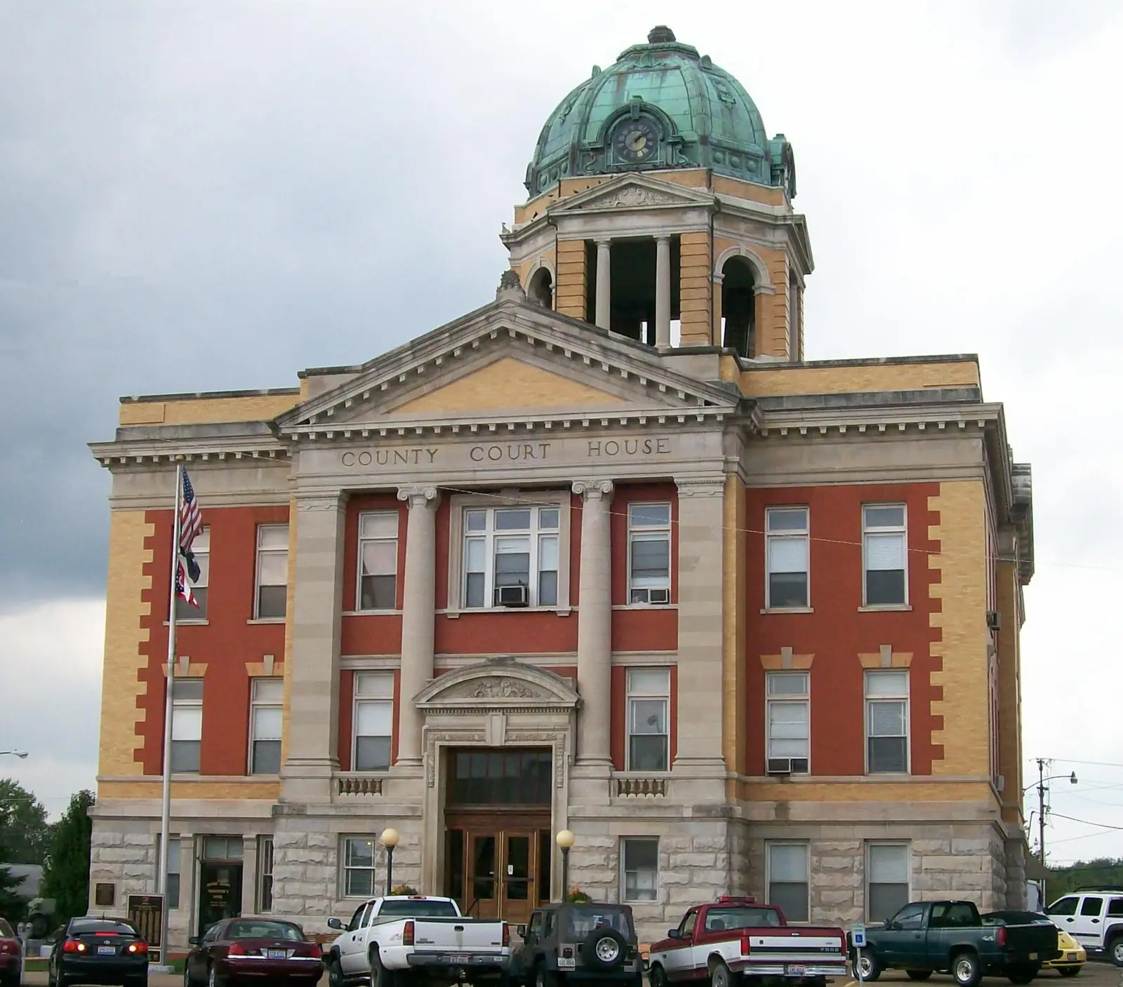 A three-story brick and stone courthouse with columns over the door and a green copper dome, with pickup trucks parked in front.
