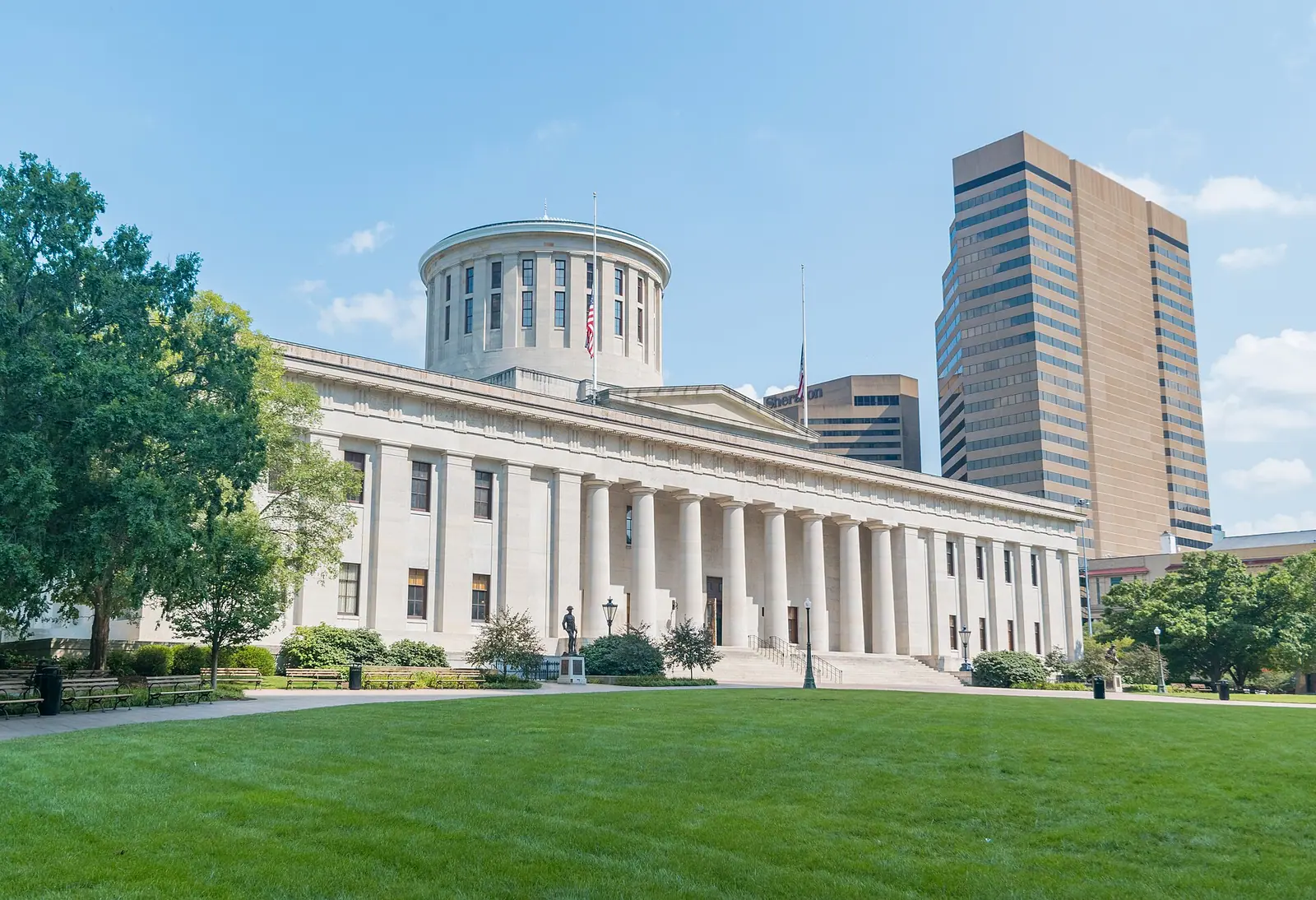 The Ohio Statehouse in Columbus, a low limestone building with a row of columns and a round drum at its center, behind a lawn.