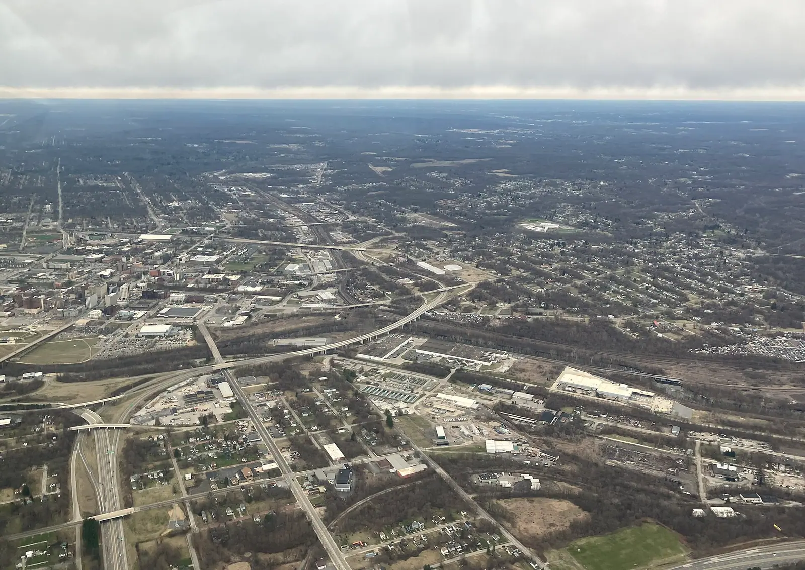 An aerial view of a small city under low cloud: a cluster of mid-rise buildings at left, highways, rail lines and industrial lots, and houses among bare trees to the horizon.