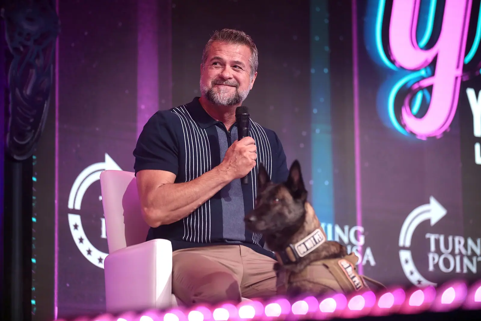Victor Marx seated on a stage lit in pink and blue, holding a microphone, with a dog in a working harness sitting beside him.